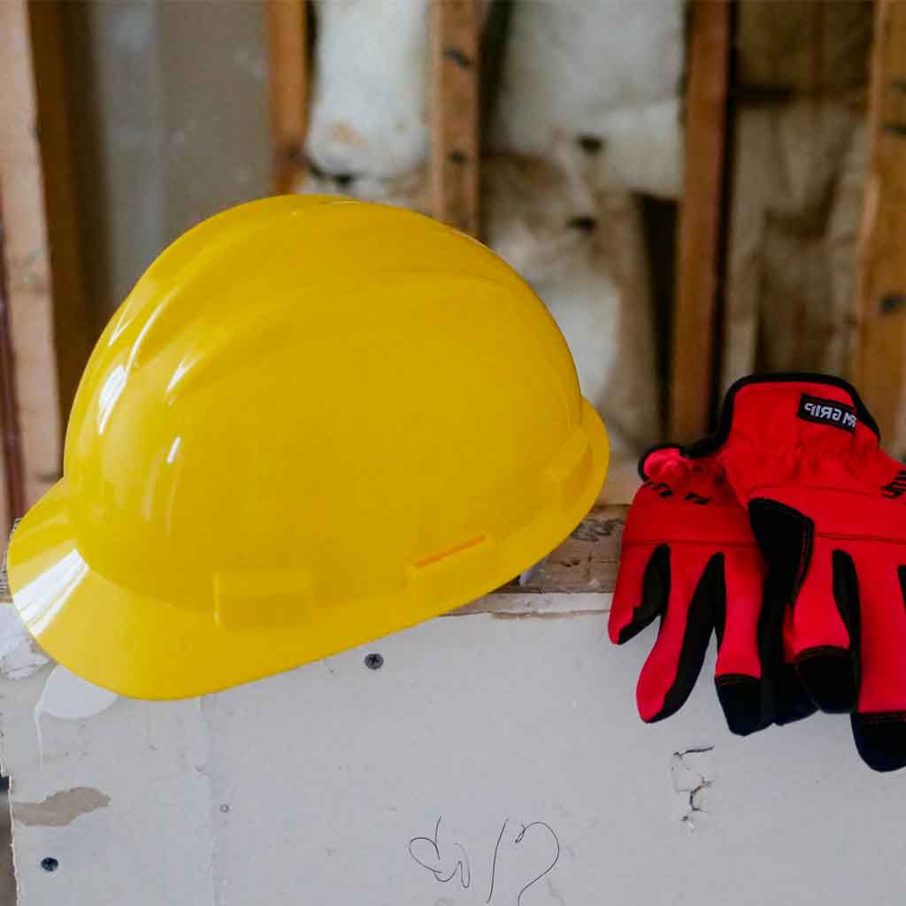 Yellow construction hard hat and red safety work gloves on construction site with exposed wooden framing and insulation, workplace safety equipment for building projects