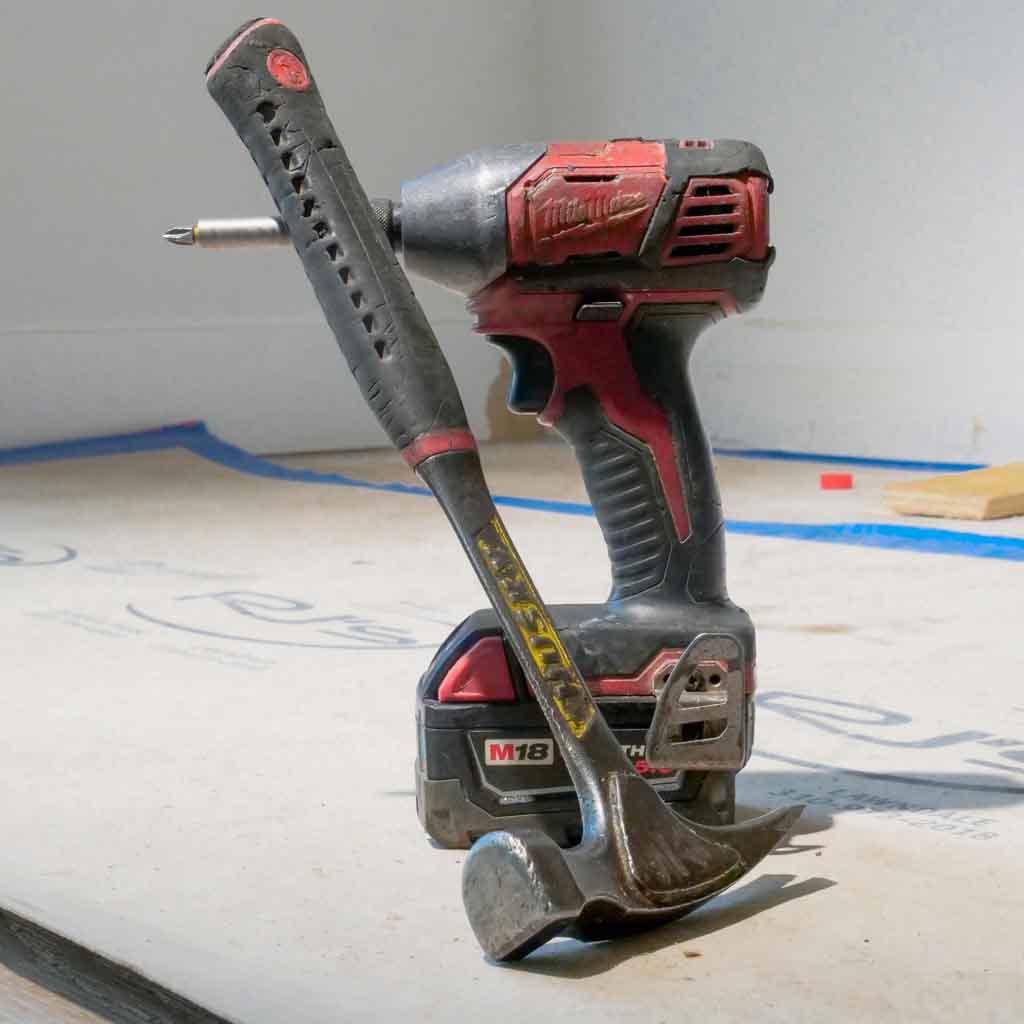 Milwaukee cordless impact driver and claw hammer construction tools on job site with blue painter's tape, essential equipment for building and renovation projects
