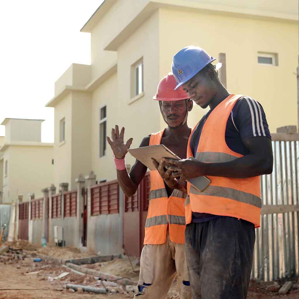 Construction workers in safety gear and hard hats reviewing building plans on tablet at residential construction site with new home development in background